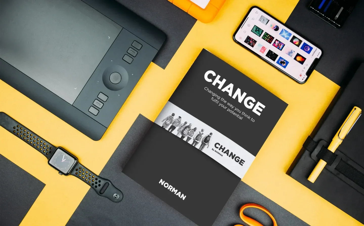 A book wrapped with a belly band on a desk, surrounded by a phone, watch, scissors and graphics tablet
