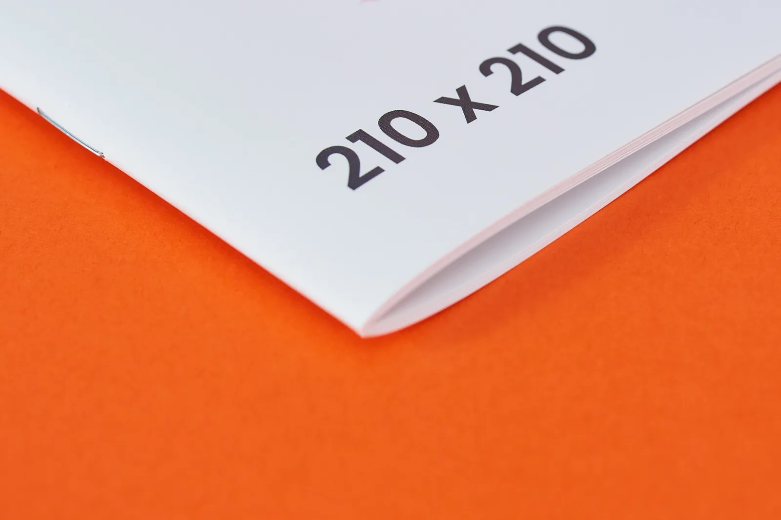 Close up of a 210 x 210mm saddle stitched stapled booklet showing the spine on a bright orange background