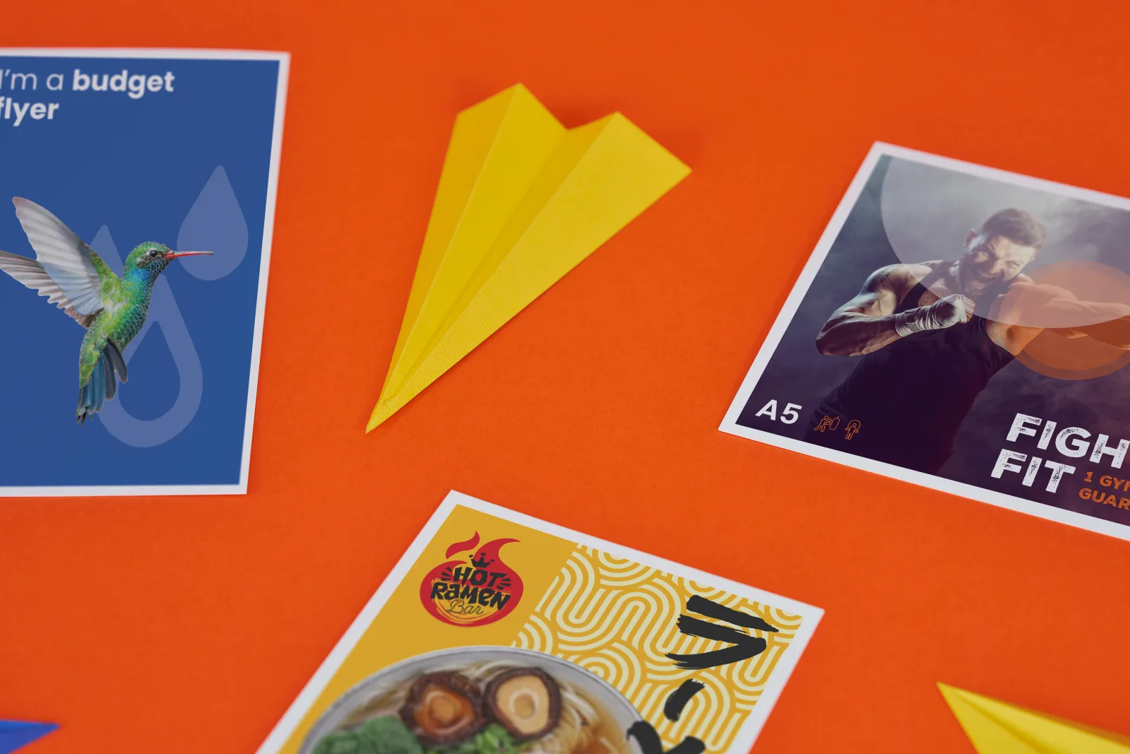 close up of 3 printed budget flyers, blue A5 portrait with hummingbird, A5 landscape with boxer and a A6 portrait yellow flyer with a bowl of ramen on a bright orange background with 2 yello