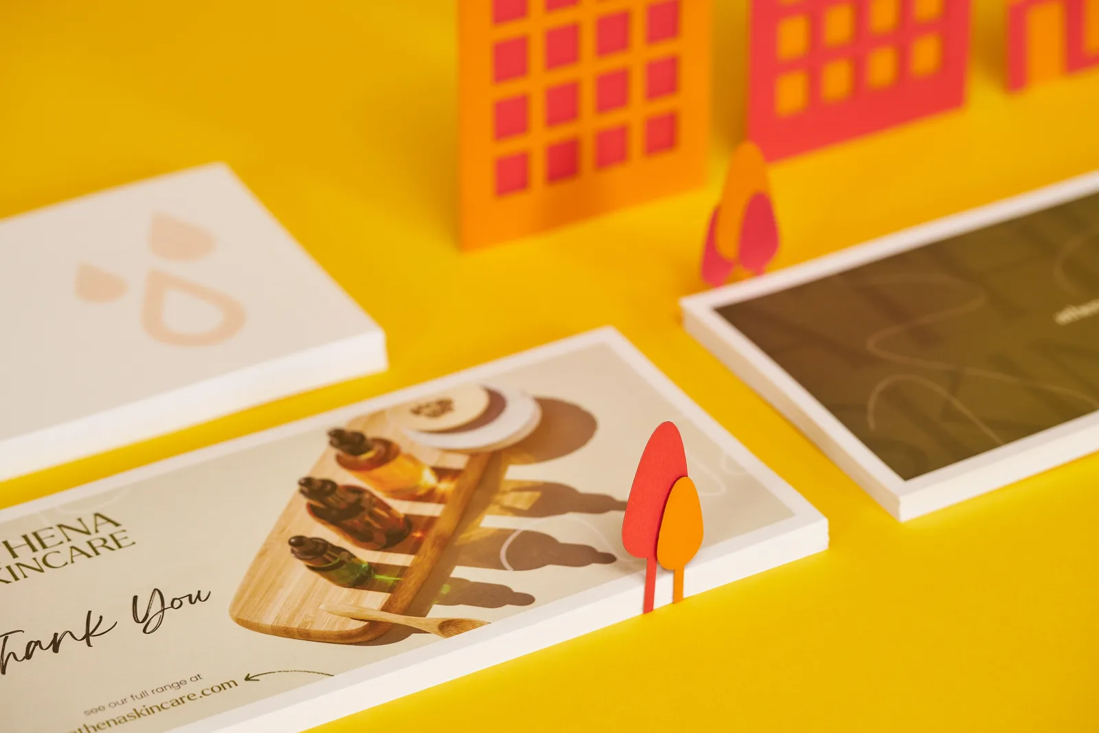 close up of 3 piles of dl printed compliment slips one saying 'athena skincare thank you' on a bright yellow background with red and orange paper buildings and trees