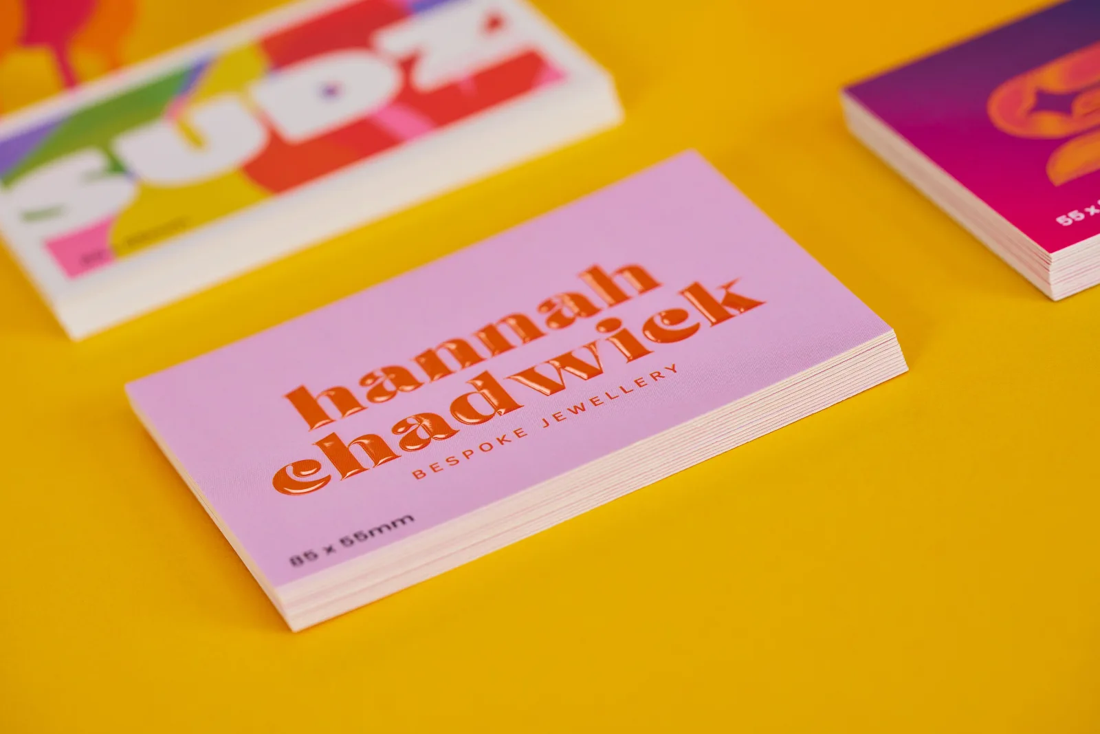 3 piles of business cards one rainbow print with text 'sudz' and one pile with spot uv text 'hannah chadwick' on a bright yellow background