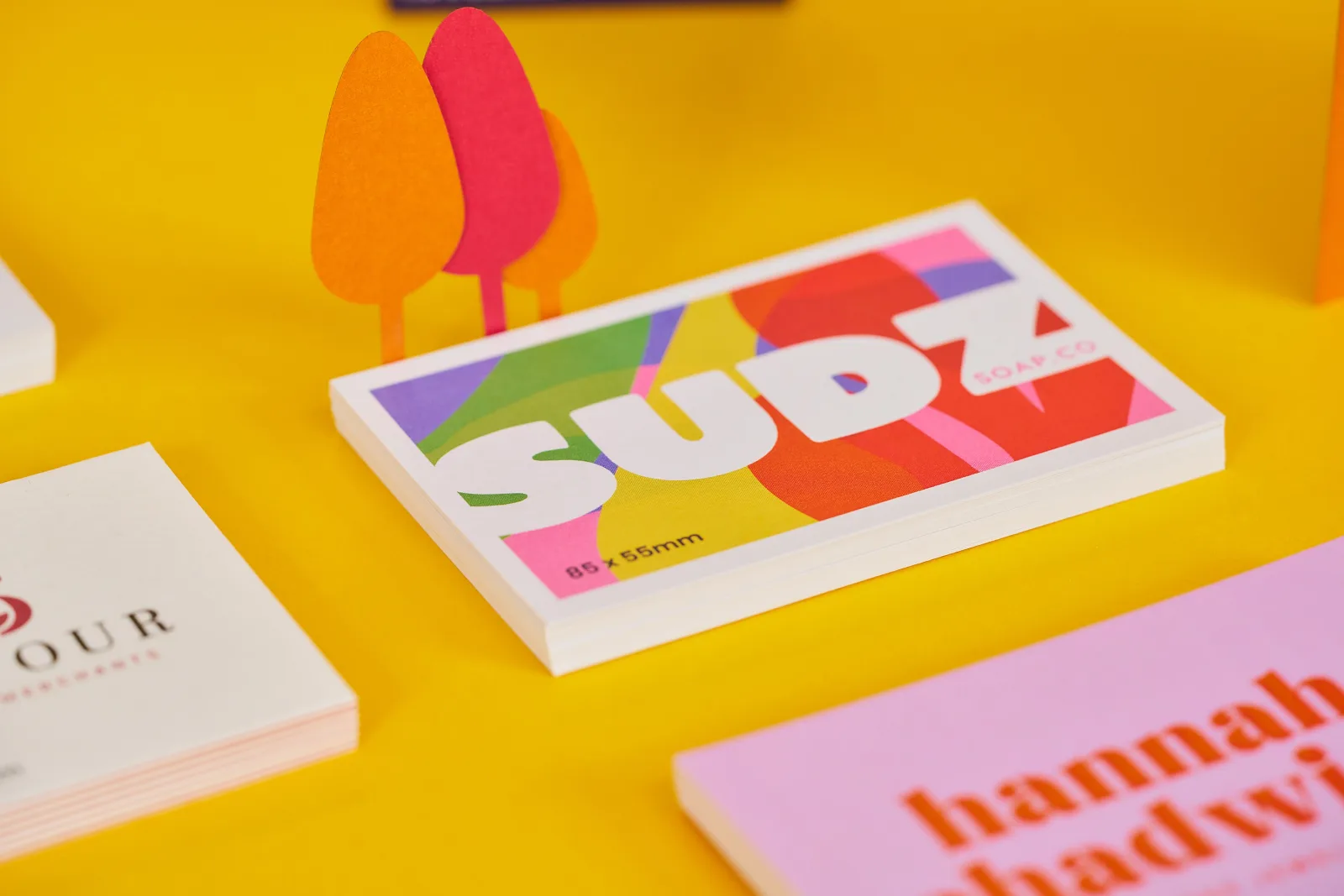 pile of 3 custom business cards with the text 'sudz' and 'hannah' with spot uv over the top on bright yellow background with red and orange paper trees