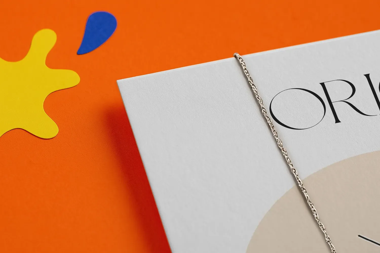 White book with a silver chain on an orange surface, beside yellow and blue paper shapes.