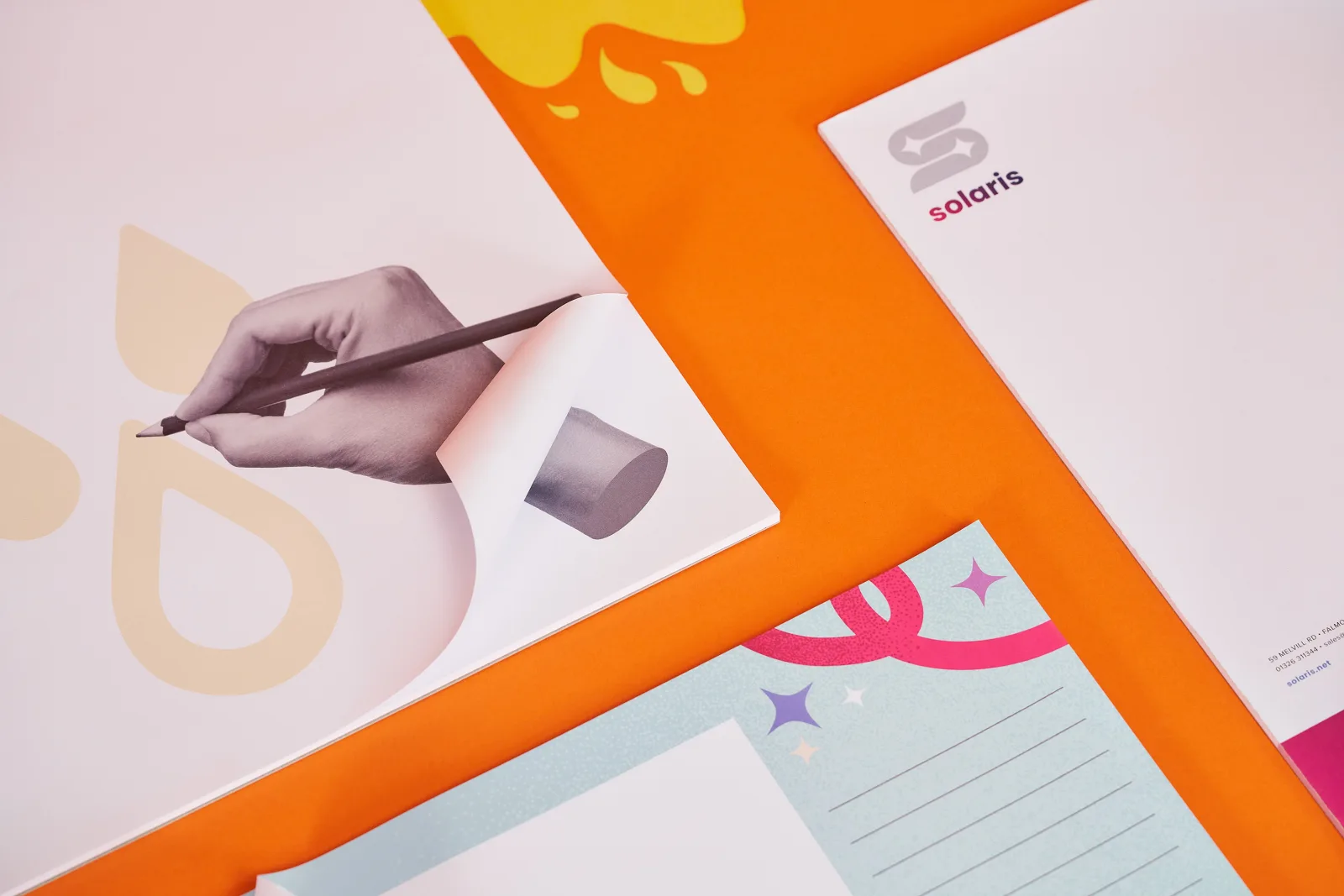 corners of 3 printed desk pads one with hand and pencil on a bright orange background with yellow paper ink spills