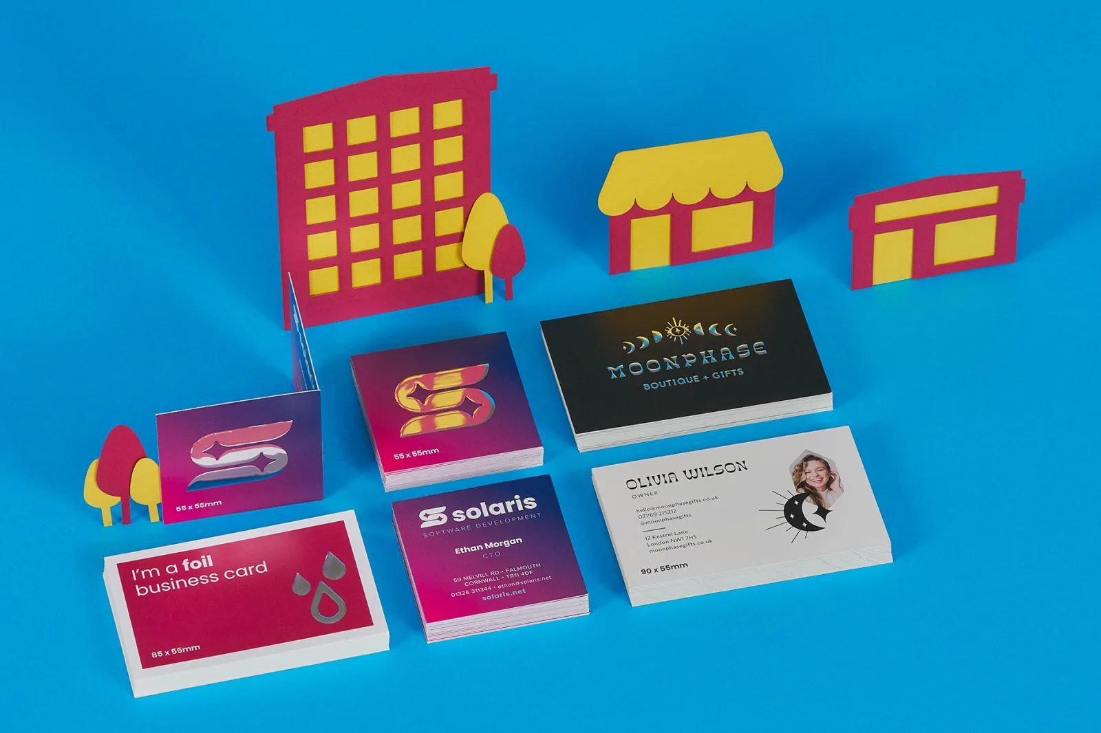 5 stacks of business cards with gold and silver foiling with text 'i'm a foil business card, solaris, moonphase' on a bright blue background with purple and yellow paper buildings and trees
