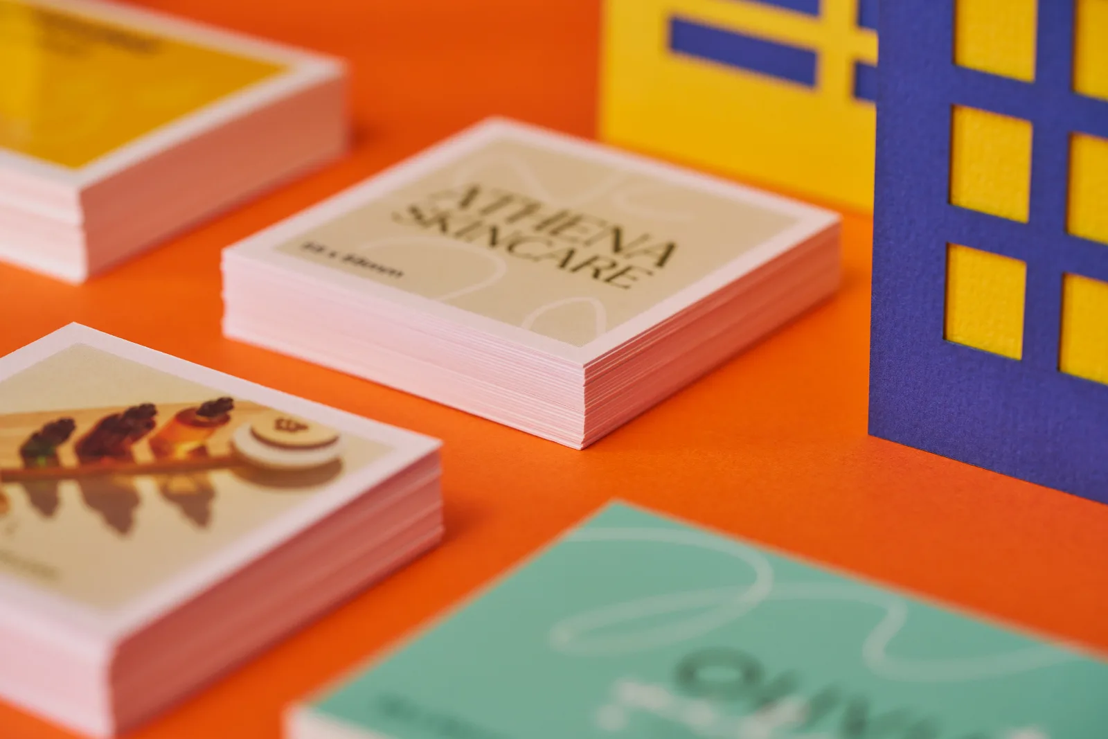 Close up of 2 piles of 55mm x 55mm cream business cards with wording ' athena skincare' on a bright orange background with yellow and blue paper buildings
