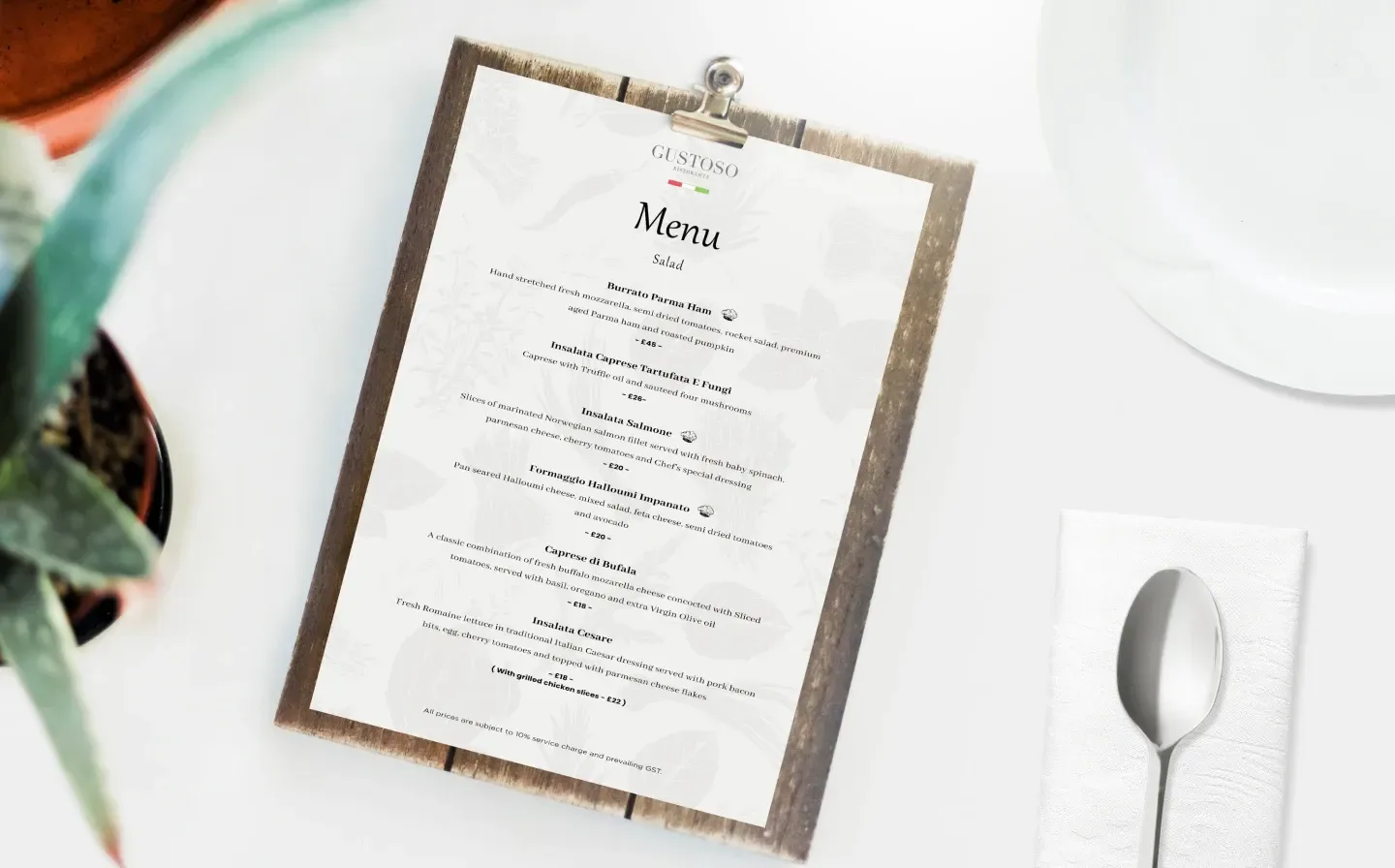 A printed menu on a clipboard, placed on a white table with a spoon, plate and a plant nearby