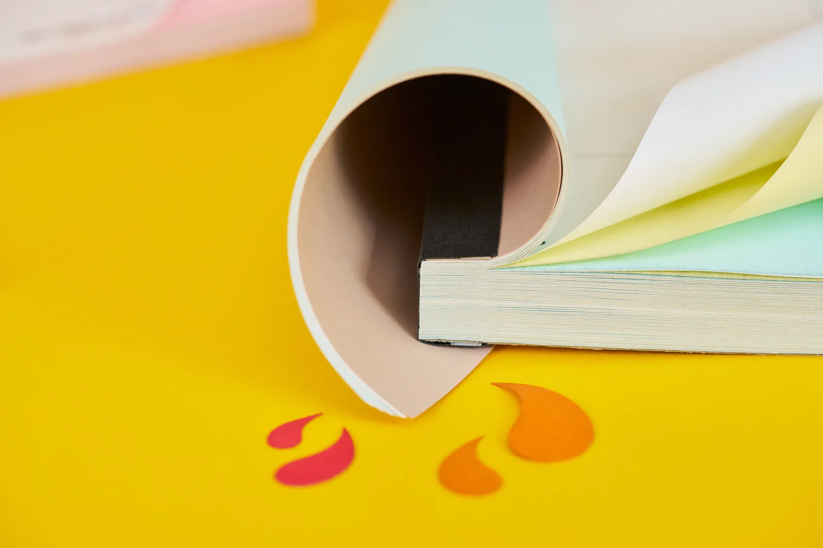 spine detail of an a5 ncr book with cover curled over showing curled white top sheet and curled yellow and blue duplicate sheets on bright yellow background with orange and red paper ink spl