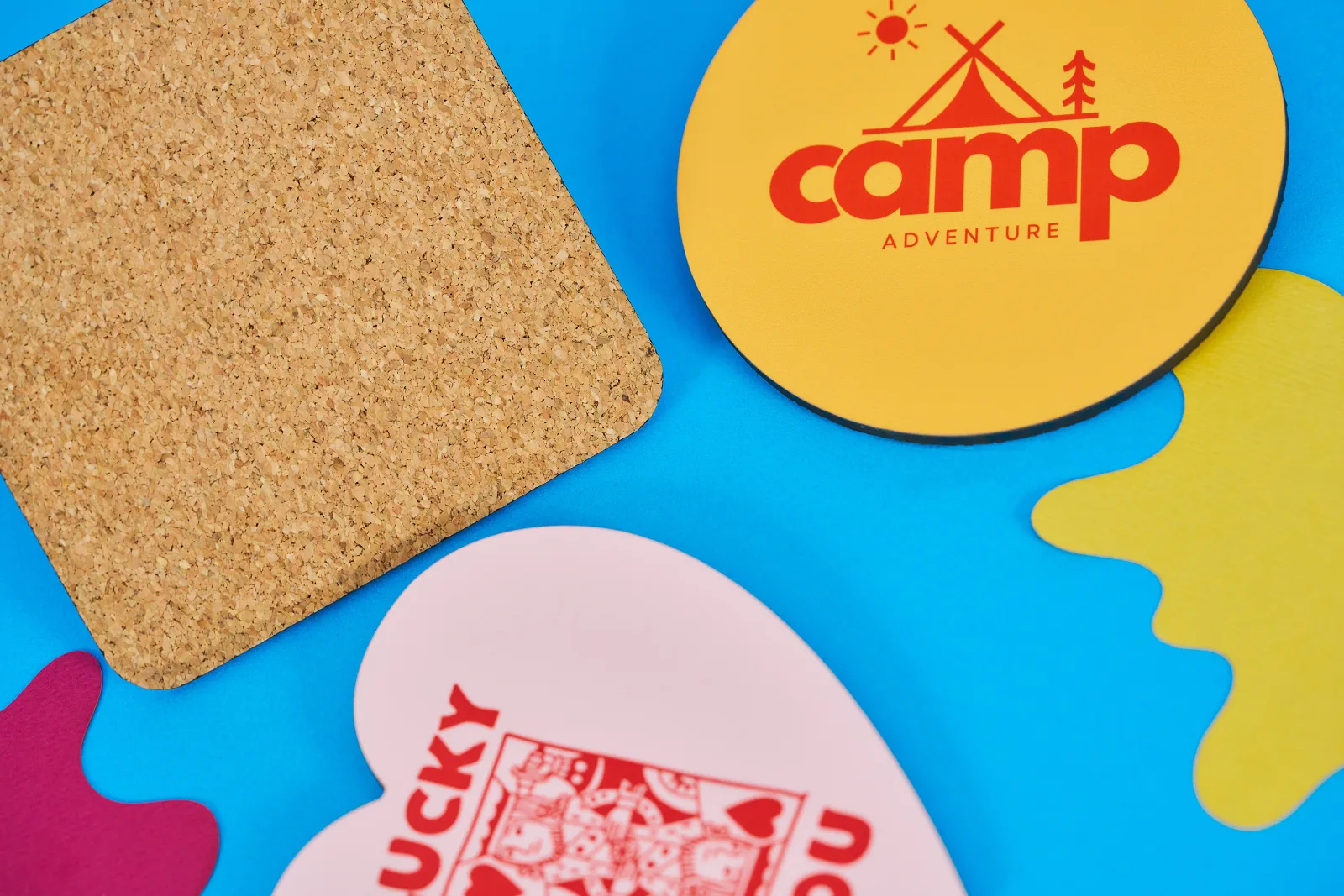yellow circle personalised coaster with text 'camp adventure' pink heart shaped coaster and the cork back of a square coaster on a bright blue background with yellow and purple paper ink spl