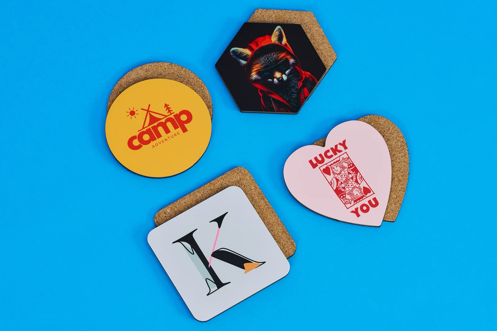 printed hexagon, square, circle and heart shaped coasters with text 'i'm a personalised coaster', 'lucky you', 'camp' with upside down coasters below showing cork back on a bright blue backg