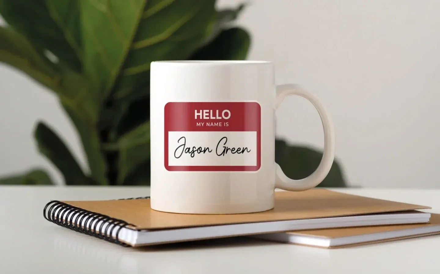  A large mug on a desk displays the name design "Hello My Name Is Jason Green," surrounded by books and a plant