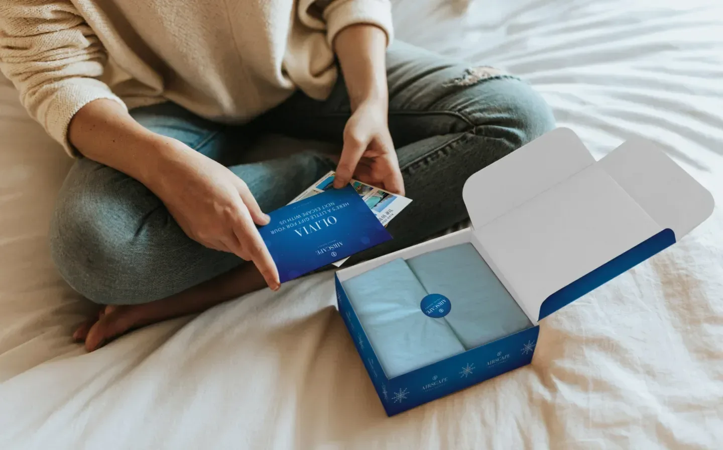 A woman sits on a bed with a blue gift box open in front of her, reading a thank you card