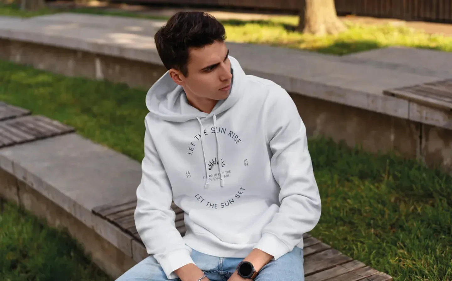 A man in a white hoodie is seated on a bench, appearing relaxed and contemplative in a serene outdoor setting