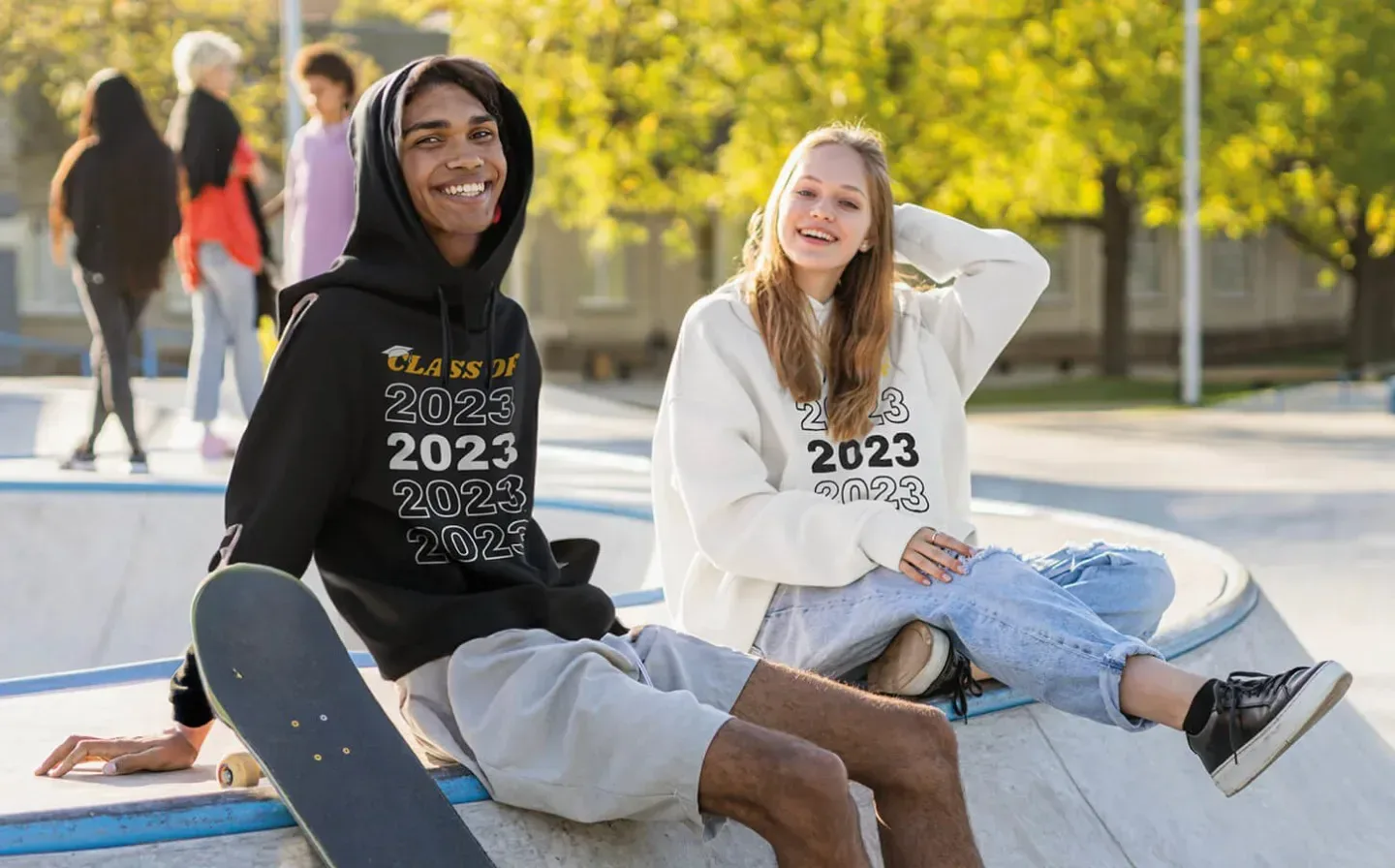 Two individuals seated on a skateboard wearing 2023 leavers shirts in front of a vibrant skate park backdrop