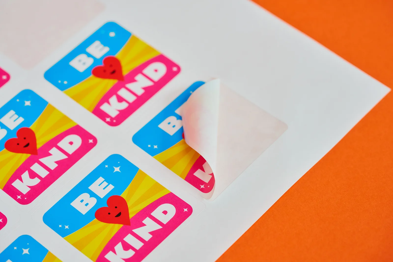 close up of a blue, yellow and pink with a red heart square sticker with rounded corners peeling off a a4 sheet with text 'be kind' on a bright orange background