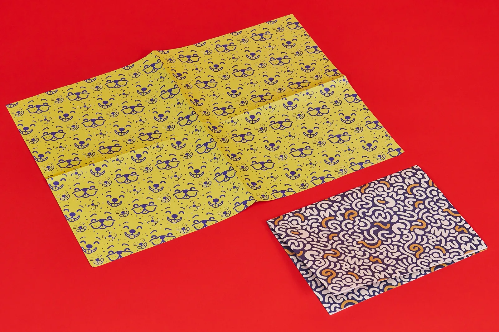 bright yellow sheet of unfolded personalised wrapping paper and a folded sheet of personalised wrapping paper with blue print on a bright red background
