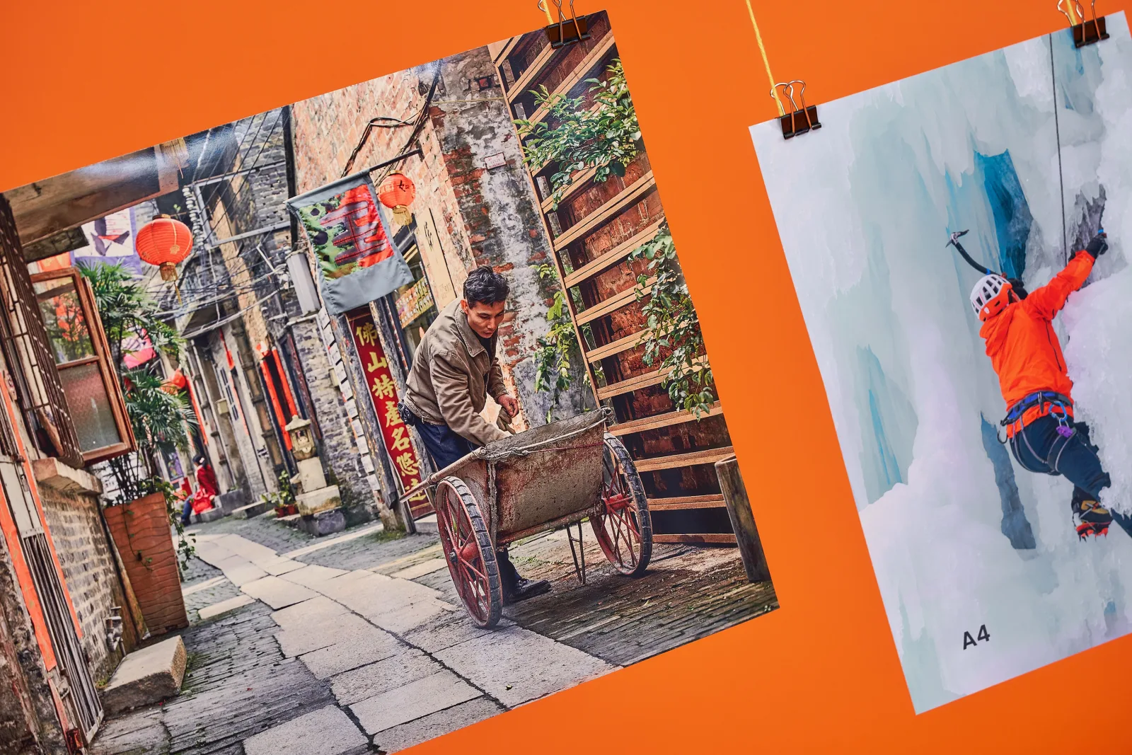 photo poster of an asian village street with a man and a wooden cart and a a4 photo poster of a man climbing ice in bright orange jacket both hanging from yellow string on a bright orange ba