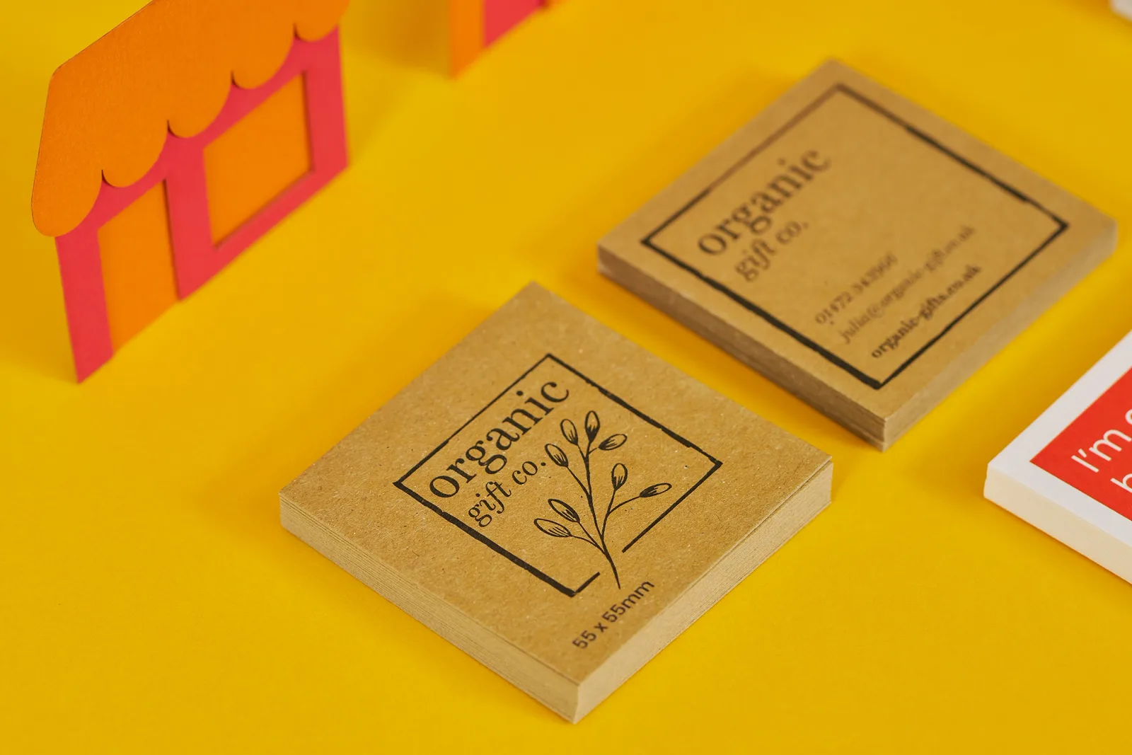 2 piles of 55 x 55mm kaft paper business cards with text 'organic gift.co' on a bright yellow background with orange and red paper building
