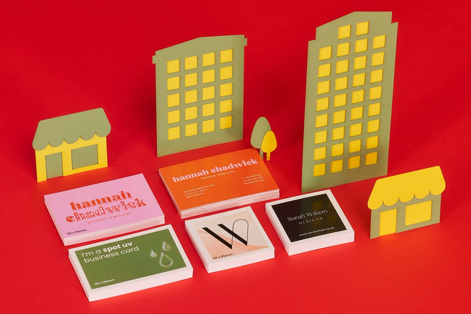 5 stacks of spot uv business cards with text 'i'm a spot uv business card' hannah chadwick' 'sarah wilson designs' on a bright red background with green and yellow buildings and trees